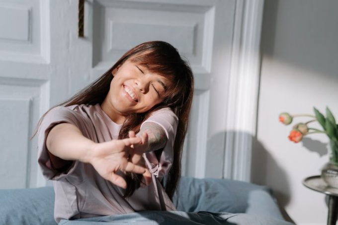 Smiling Asian woman stretching in bed, enjoying soft morning light indoors.